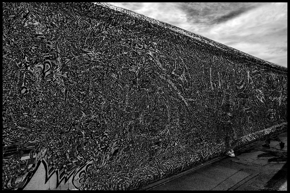 Two people appear to simply penetrate the Berlin Wall by walking through it. The artistic black and white photo shows painted fragments of the Berlin Wall with semi-transparent people.