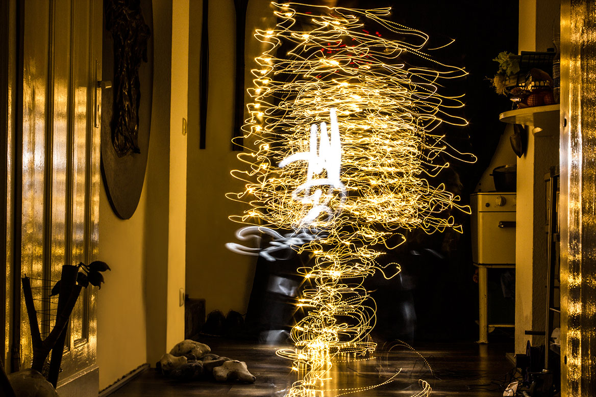Fotografische Licht-Inszenierung, Einstellung 6. Eine Frau erstrahlt durch Lichtspuren, die durch Langzeitbelichtung die Körperbewegungen nachzeichnen. Die Lichtspuren sind überwiegend in gelben Farben gehalten, pointiert durch weisse und rote Akzente. Der Körper scheint zu vibrieren.