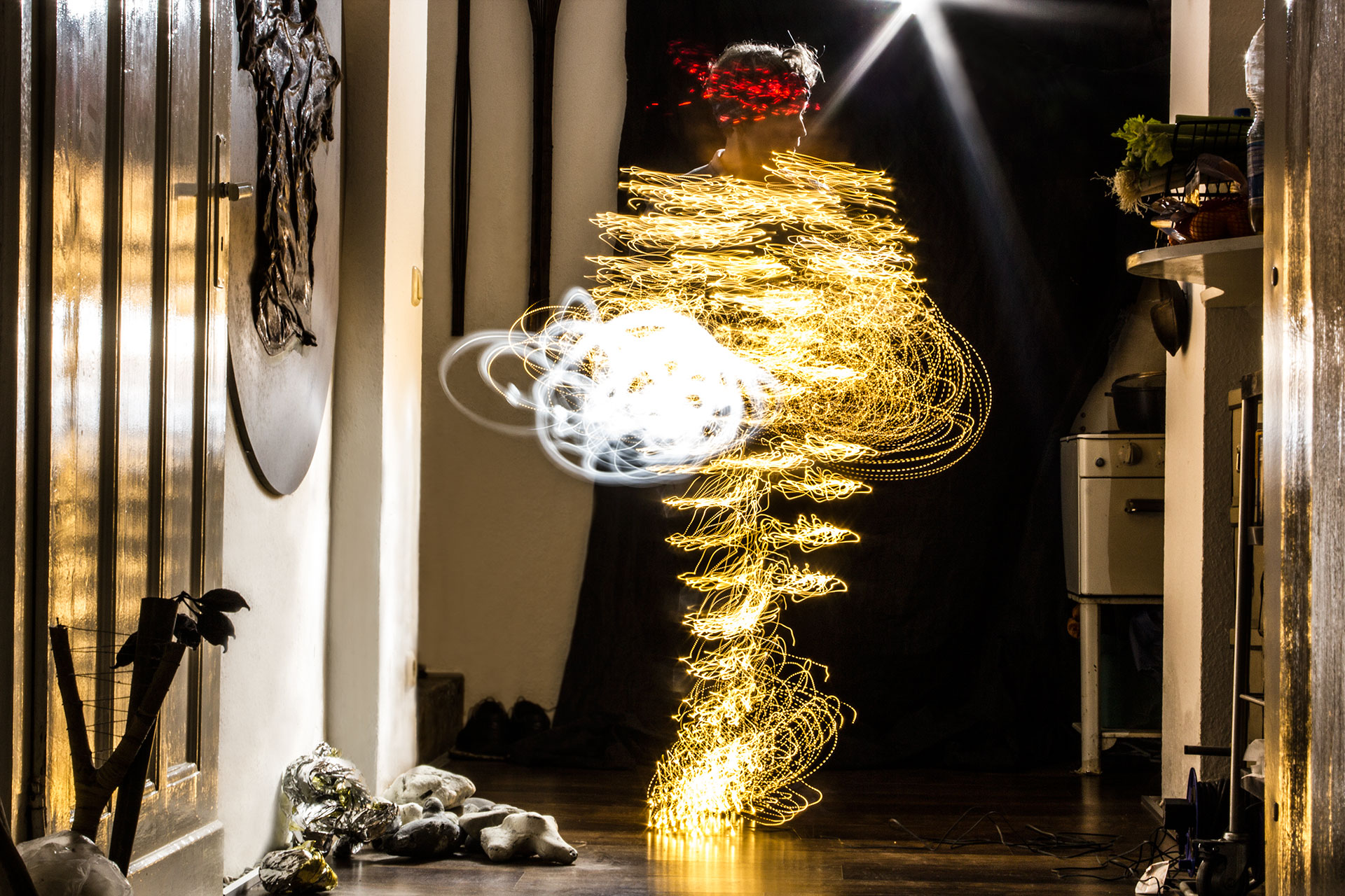 Fotografische Licht-Inszenierung, Einstellung 1. Eine Frau erstrahlt durch Lichtspuren, die durch Langzeitbelichtung die Körperbewegungen nachzeichnen. Die Lichtspuren sind überwiegend in gelben Farben gehalten, pointiert durch weisse und rote Akzente. Die Kulisse spiegelt eine Wohnung mit ausgestellten Kunstwerken.