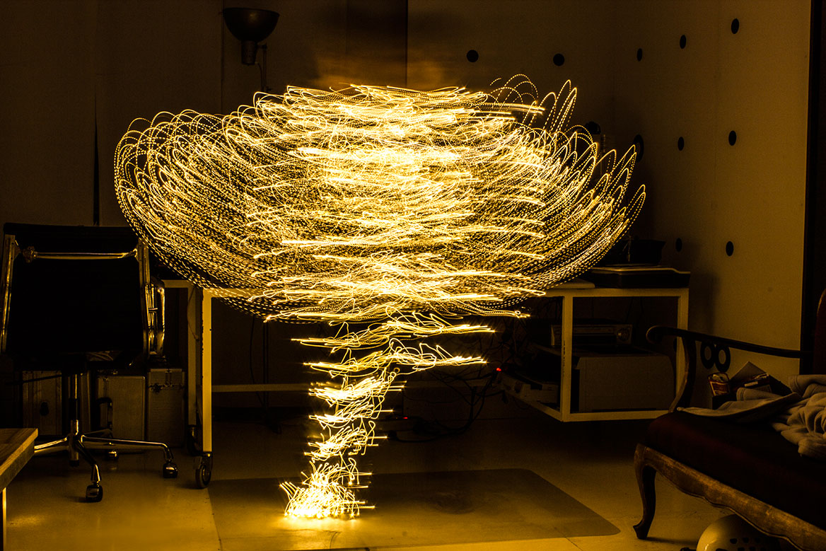 Fotografische Licht-Inszenierung, Einstellung 5. Eine Frau erstrahlt durch Lichtspuren, die durch Langzeitbelichtung die Körperbewegungen nachzeichnen. Die Langzeitbelichtung zeigt fast symmetrische Lichtspuren.