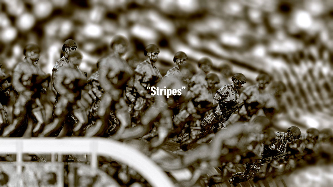 Standbild aus der Animation. Halbtotale auf die Soldatenreihen, von der Seite gesehen. Das englische Wort -Stripes- steht in weißer Typografie zentral im Bild und ist mit An- und Abführungszeichen gesetzt. Darunter ist das mathematische Unendlichkeitszeichen abgebildet, dass in der Animation fast durchweg um eine unsichtbare, vertikale Achse rotiert.