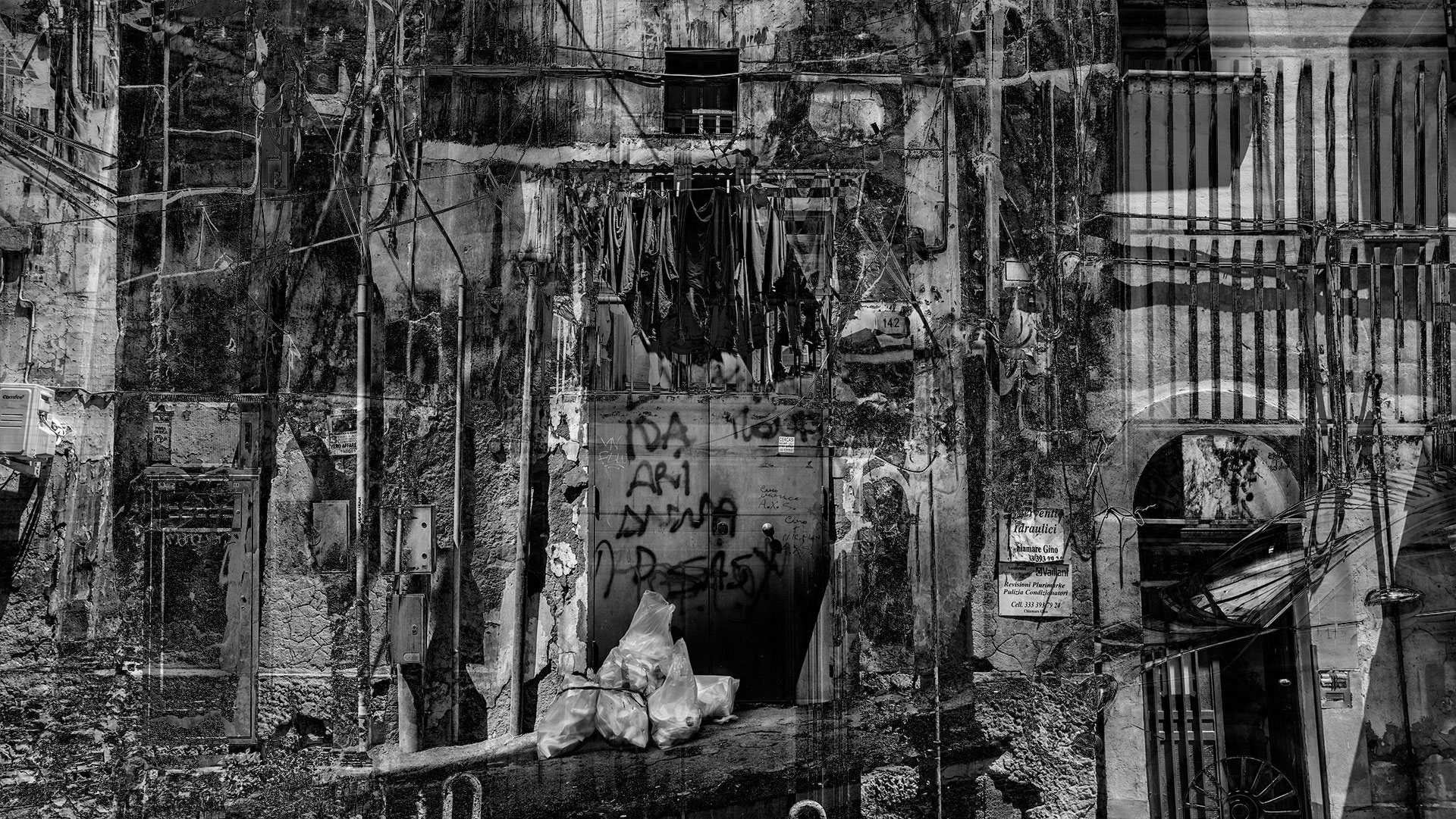 Laundry hung up outside, on the upper floor above an entrance area. A front door, downspouts and scaffolding elements, a lattice fence, filled garbage bags in the entrance area and chipped facade plaster again create a gloomy pictorial mood.